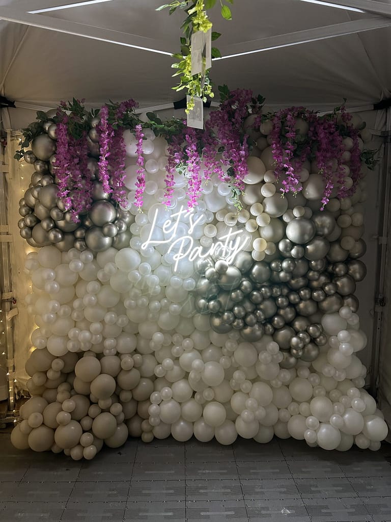 White, silver and grey balloon wall with purple floral garland and 'Let's Party' neon sign under marquee